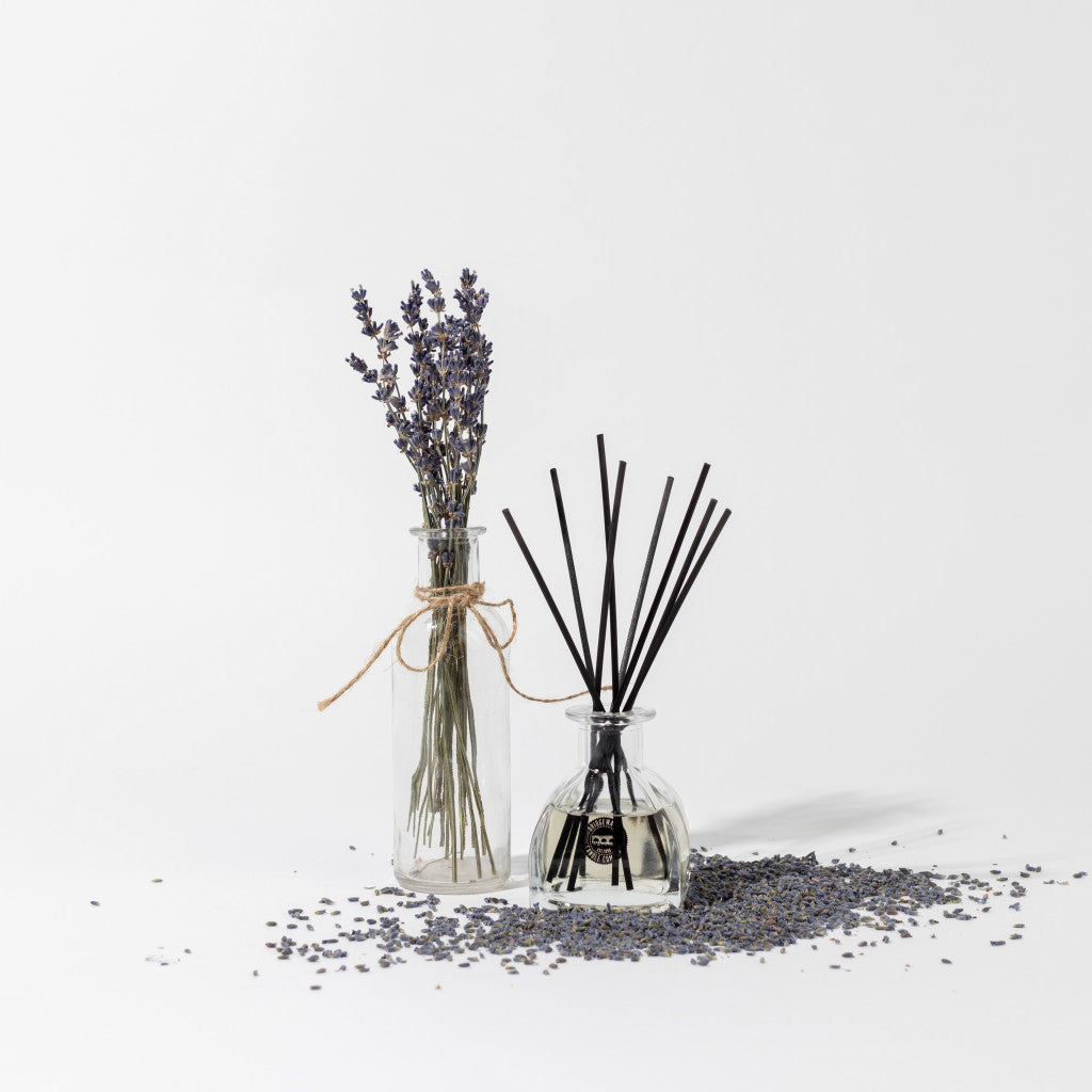 A glass bottle with lavender stems tied with twine sits beside a Bridgewater Candle Company Lavender Fields Reed Diffuser with fiber reeds, surrounded by dried lavender on a white background.