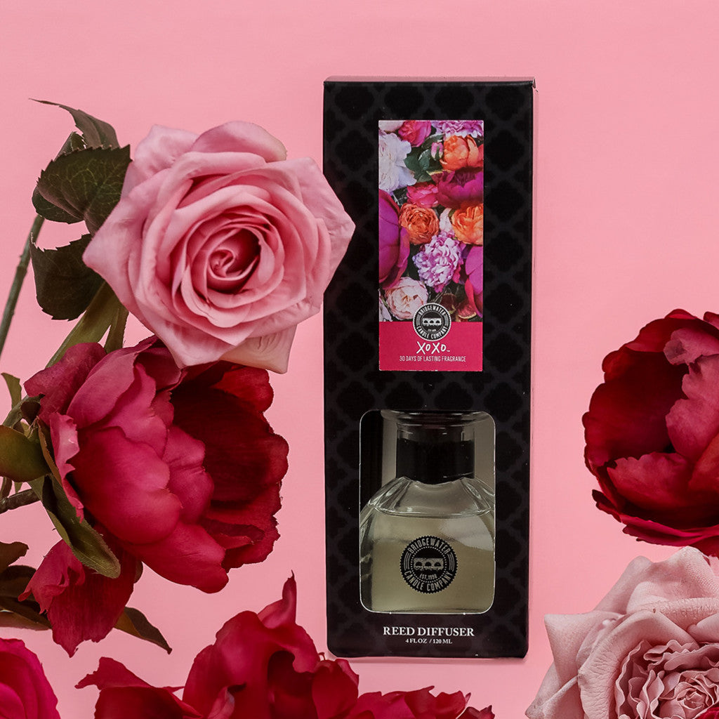 The Bridgewater Candle Company XOXO Reed Diffuser comes in a black box with colorful flowers, surrounded by pink and red roses on a light pink background. This premium diffuser uses fiber reeds for a long-lasting floral scent.