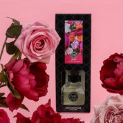 The Bridgewater Candle Company XOXO Reed Diffuser comes in a black box with colorful flowers, surrounded by pink and red roses on a light pink background. This premium diffuser uses fiber reeds for a long-lasting floral scent.