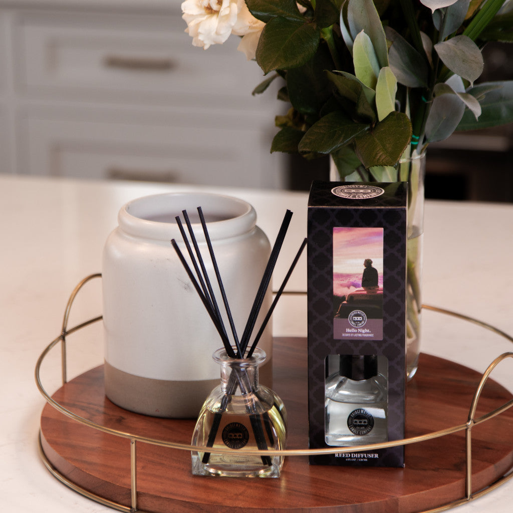 A Bridgewater Candle Company Hello Night Reed Diffuser with fiber reeds stands by its box on a round wooden tray. A large ceramic container and a vase of white flowers and green leaves complete the elegant kitchen counter arrangement.