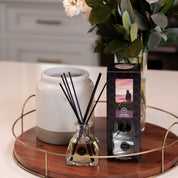 A Bridgewater Candle Company Hello Night Reed Diffuser with fiber reeds stands by its box on a round wooden tray. A large ceramic container and a vase of white flowers and green leaves complete the elegant kitchen counter arrangement.