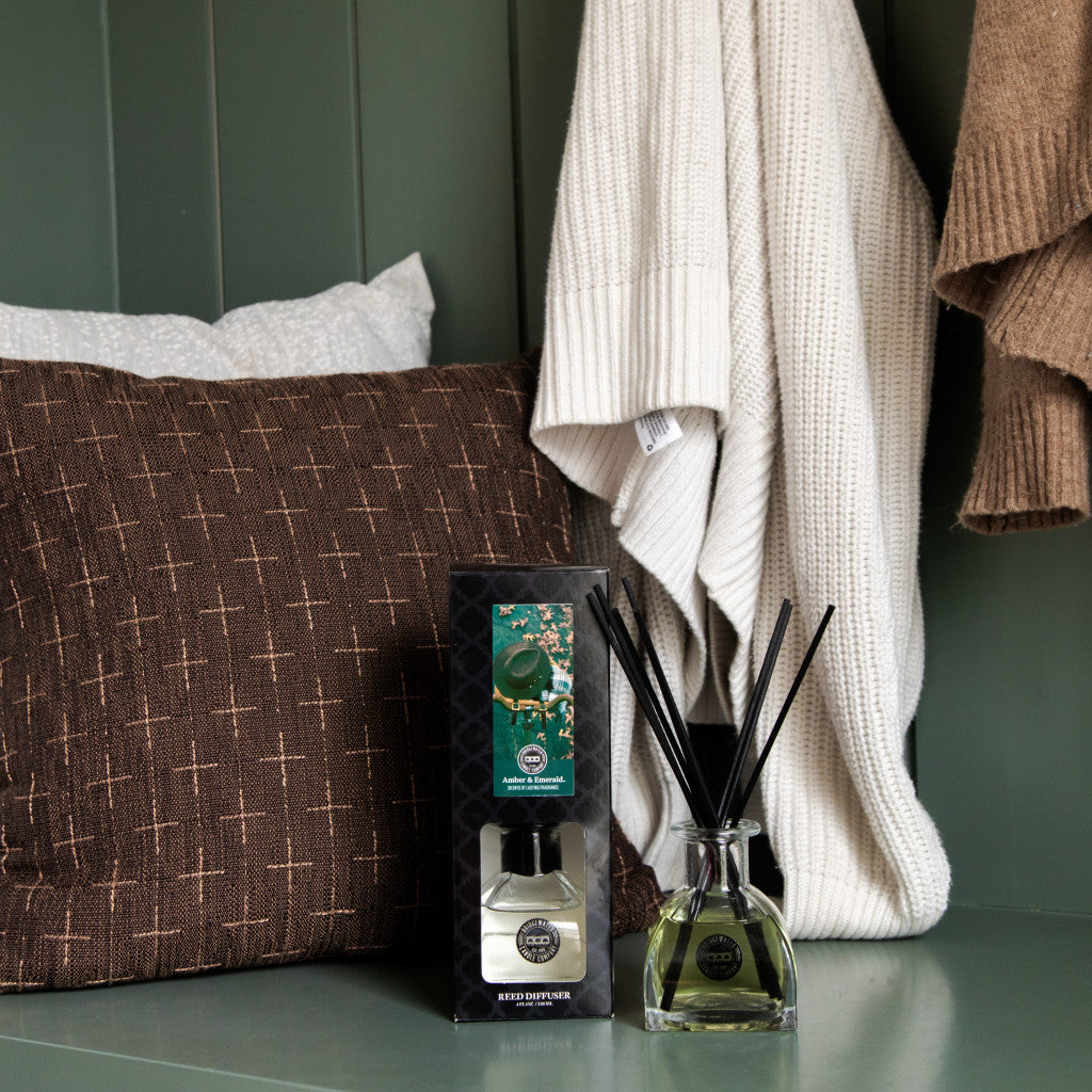 A Bridgewater Candle Company Amber & Emerald Reed Diffuser sits on a green shelf beside a brown cushion and white bedding, filling the room with fragrance as cream and brown sweaters hang on hooks above.