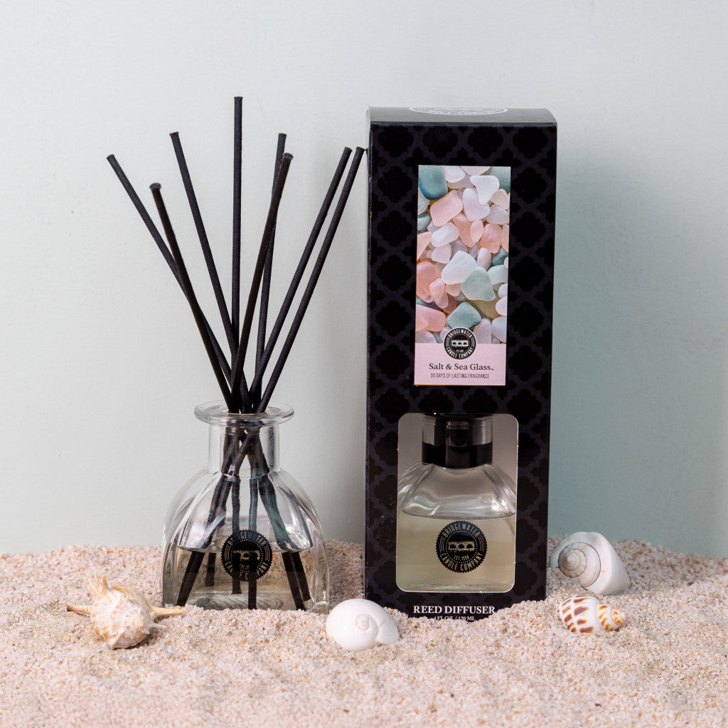 The Bridgewater Candle Company Salt & Sea Glass Reed Diffuser, featuring black reeds in a glass bottle and a pastel stone-adorned box, rests on sand with seashells—ideal for enhancing your homes fragrance ambiance.