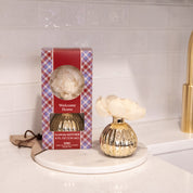 A boxed Welcome Home Mercury Flower Diffuser by Bridgewater Candle Company and a gold vase with a cream flower sit on a white counter near a sink with a gold faucet; the box has a red plaid design and brings elegant fragrance to your home.