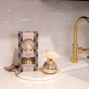 The Bridgewater Candle Company Afternoon Retreat Mercury Flower Diffuser, featuring a white flower, is displayed on a round marble tray with its packaging, adding home fragrance to a bright kitchen with gold accents and white tile backsplash.