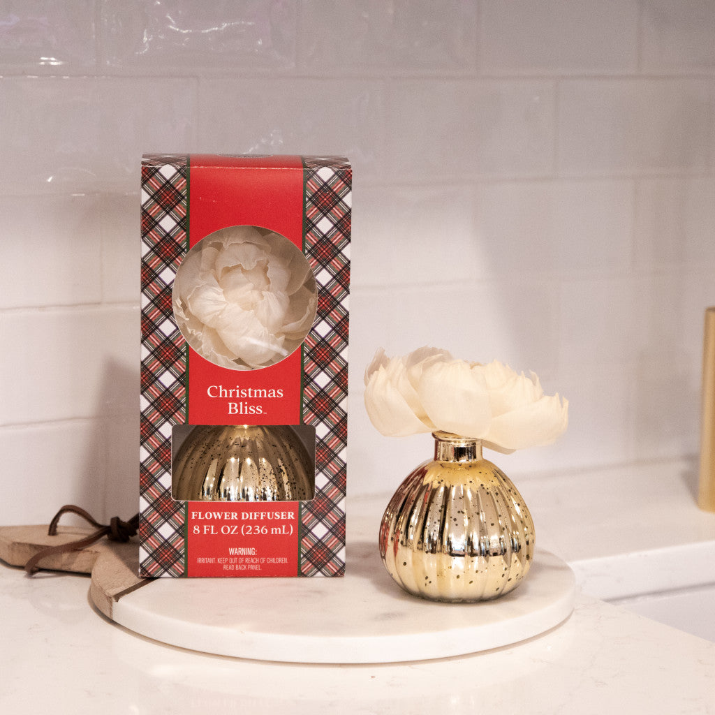 A boxed Bridgewater Candle Company Christmas Bliss Holiday Mercury Flower Diffuser sits beside an open gold and white diffuser with a large cream flower, displayed on a white round tray against a white tile background.