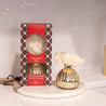 A boxed Bridgewater Candle Company Christmas Bliss Holiday Mercury Flower Diffuser sits beside an open gold and white diffuser with a large cream flower, displayed on a white round tray against a white tile background.