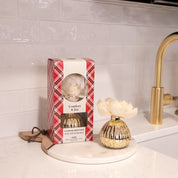 The Bridgewater Candle Company Comfort & Joy Holiday Mercury Flower Diffuser sits boxed on a kitchen counter beside a gold vase diffuser with a white flower, near a gold faucet and white tile backsplash.