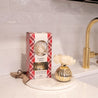 The Bridgewater Candle Company Comfort & Joy Holiday Mercury Flower Diffuser sits boxed on a kitchen counter beside a gold vase diffuser with a white flower, near a gold faucet and white tile backsplash.