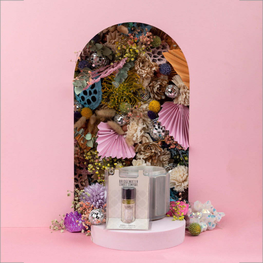 The Sweet Grace Home Fragrance Oil by Bridgewater Candle Company is displayed with a glass holder in front of a pink arch decorated with bright flowers, foliage, paper fans, and disco balls—setting a dreamy mood for your scent experience.