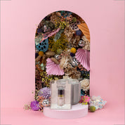The Sweet Grace Home Fragrance Oil by Bridgewater Candle Company is displayed with a glass holder in front of a pink arch decorated with bright flowers, foliage, paper fans, and disco balls—setting a dreamy mood for your scent experience.