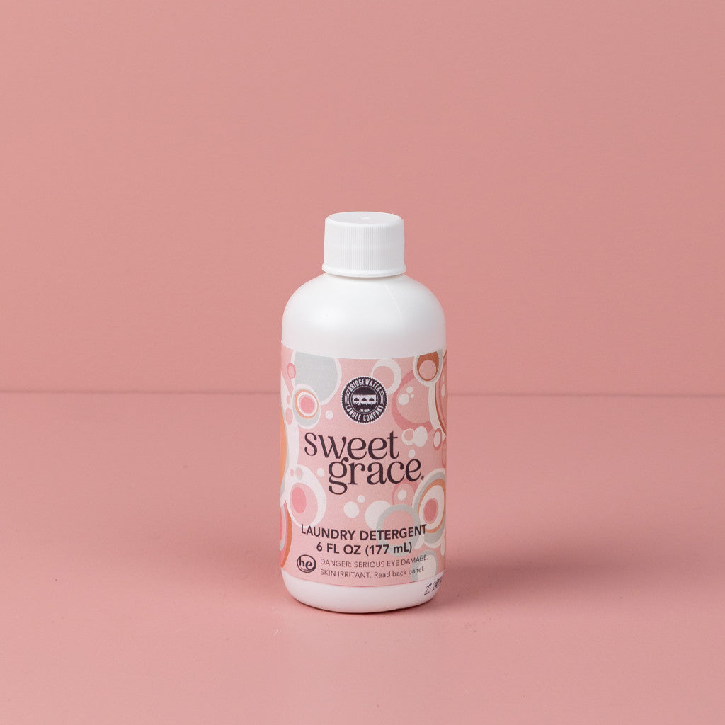 A 6oz bottle of Sweet Grace Laundry Detergent by Bridgewater Candle Company stands upright on a pink surface, showcasing its pink and white patterned label with decorative circles, fabric-safe formula, and long-lasting scent.