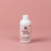 A 6oz bottle of Sweet Grace Laundry Detergent by Bridgewater Candle Company stands upright on a pink surface, showcasing its pink and white patterned label with decorative circles, fabric-safe formula, and long-lasting scent.