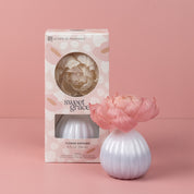 The Bridgewater Candle Company Sweet Grace Flower Diffuser, featuring a white ceramic base with a pink flower and Sweet Grace fragrance, is displayed before pastel-pink floral window packaging, perfectly matching the soft pink background.
