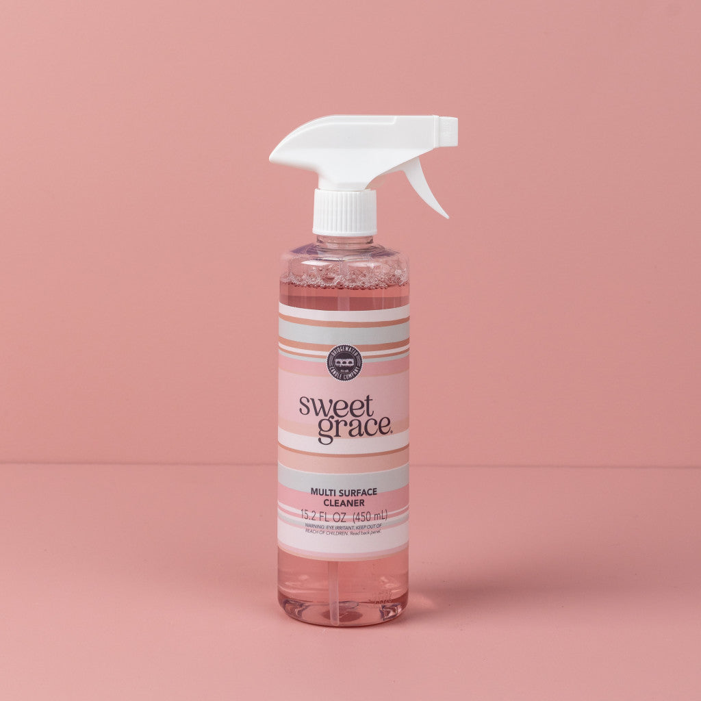 A pink spray bottle of Bridgewater Candle Company Sweet Grace Multi-Surface Cleaner with a white nozzle stands against a pink background. Its white and pink striped label highlights the signature Sweet Grace fragrance.