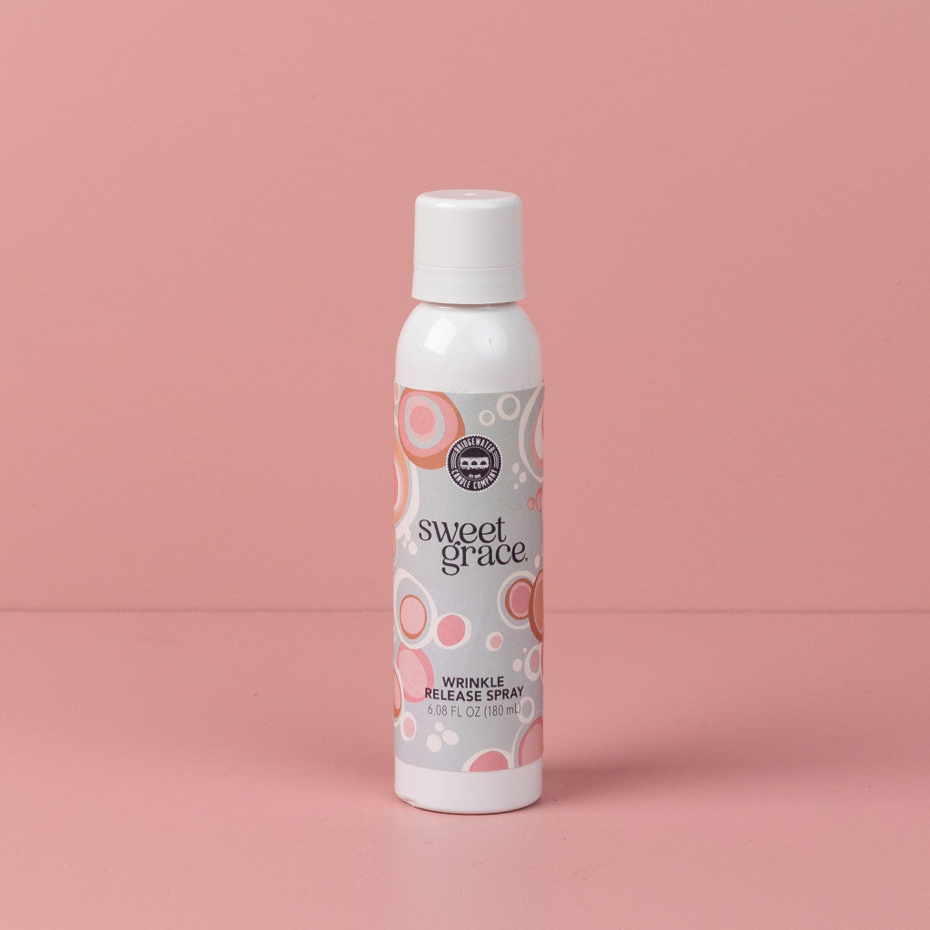 A white Bridgewater Candle Company Sweet Grace Wrinkle Release Spray bottle stands upright on a pink surface with a pink background. The label displays pink and orange circular patterns.