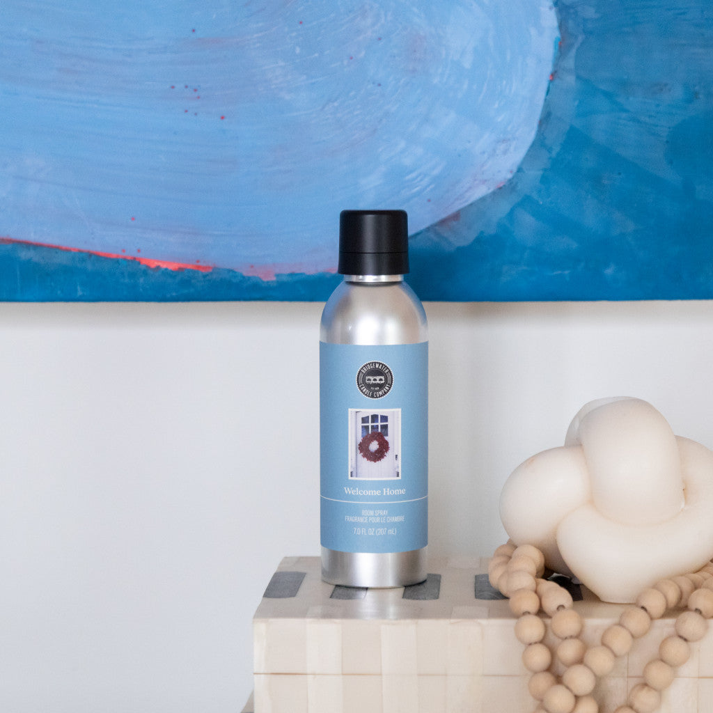 Bridgewater Candle Companys Welcome Home Non-Aerosol Room Spray with a blue label sits on a checkered box beside a knotted cream-colored candle and wooden beads, with a blue abstract painting in the background.