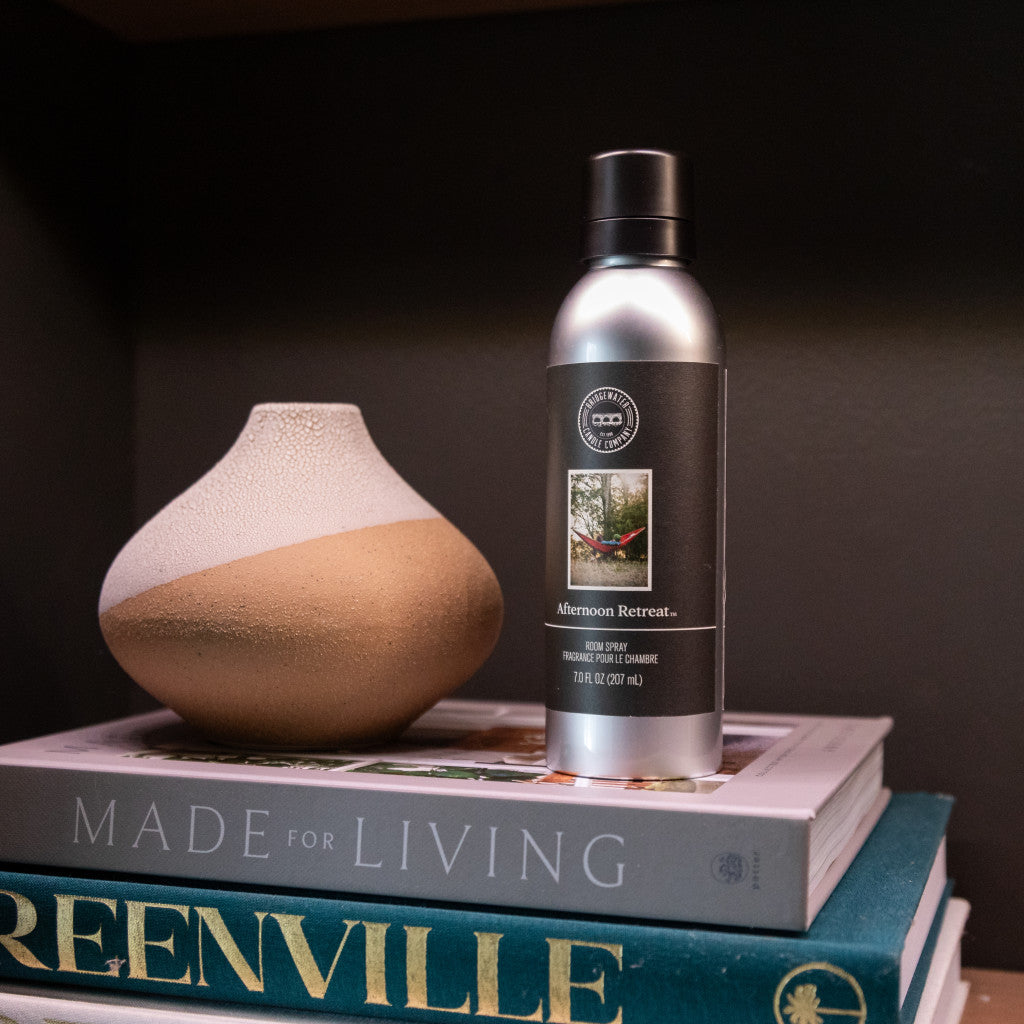A small, round ceramic vase and a Bridgewater Candle Company Afternoon Retreat Non-Aerosol Room Spray sit atop two stacked books, MADE FOR LIVING and GREENVILLE, set against a dark background.