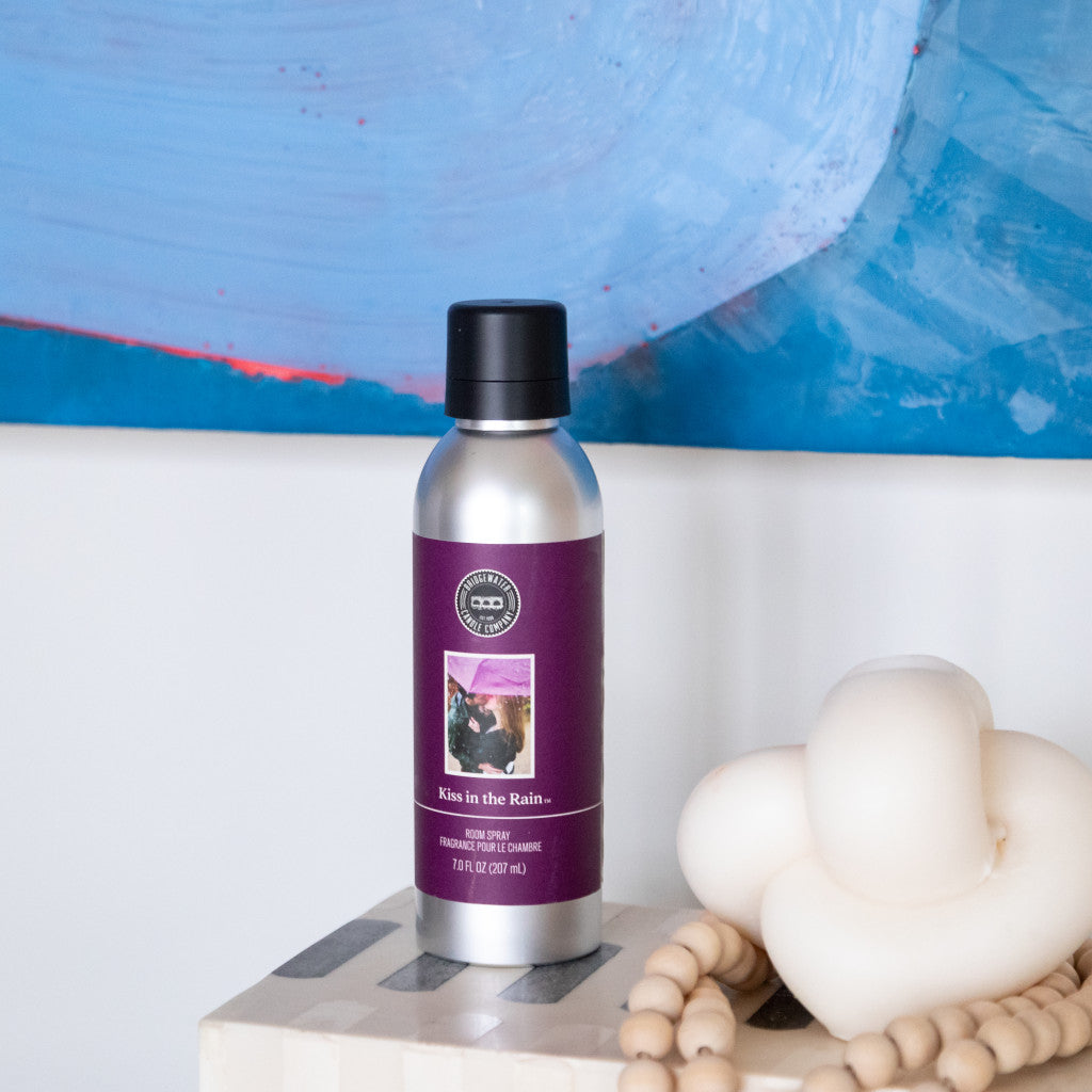 The Kiss in the Rain Non-Aerosol Room Spray by Bridgewater Candle Company, in a silver bottle with a purple label, adds elegance to any space—perfectly styled beside decor like a beige knotted sculpture and wooden beads.