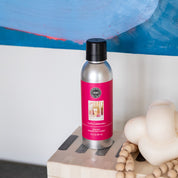 A silver bottle of Bridgewater Candle Company’s Let’s Celebrate Non-Aerosol Room Spray with a pink label sits beside a cream sculptural candle and wooden beads, with an abstract blue painting—ideal for elevating your home fragrance.