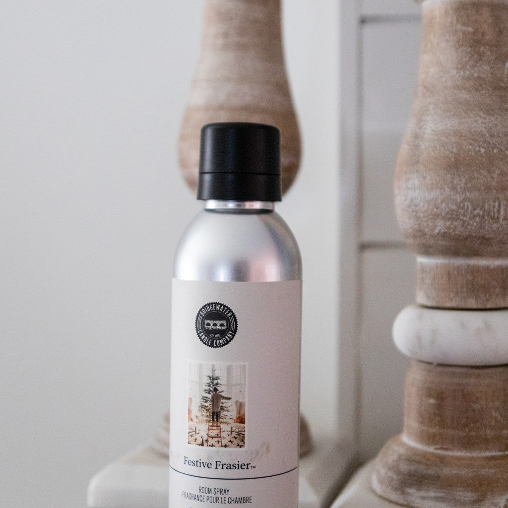 A metallic Festive Frasier Non-Aerosol Room Spray by Bridgewater Candle Company sits on a white surface, with wood decor behind it. The label shows a decorated Christmas tree, adding an eco-friendly, festive touch to your home fragrance.