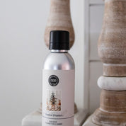 A metallic Festive Frasier Non-Aerosol Room Spray by Bridgewater Candle Company sits on a white surface, with wood decor behind it. The label shows a decorated Christmas tree, adding an eco-friendly, festive touch to your home fragrance.