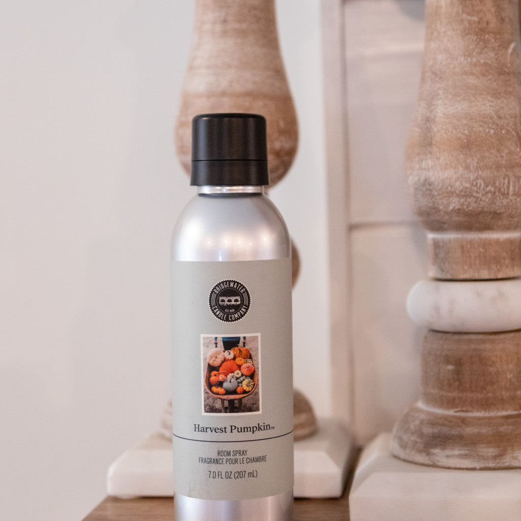 A silver can of Bridgewater Candle Company’s Harvest Pumpkin Non-Aerosol Room Spray with a black cap sits on wood, its label decorated with pumpkins and autumn decor, while wooden pillars in the background add an eco-friendly touch.