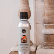 A silver can of Bridgewater Candle Company’s Harvest Pumpkin Non-Aerosol Room Spray with a black cap sits on wood, its label decorated with pumpkins and autumn decor, while wooden pillars in the background add an eco-friendly touch.