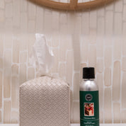 A white tissue box with a raised geometric pattern sits next to a Bridgewater Candle Company Christmas Bliss Non-Aerosol Room Spray can on a bathroom counter against a tiled wall.