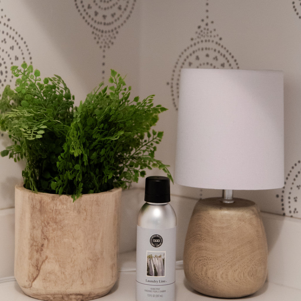 A small wooden lamp with a white shade, a potted green plant in a light wooden pot, and Bridgewater Candle Companys Laundry Line Non-Aerosol Room Spray sit on a white shelf against a patterned wall.