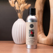 A silver Comfort & Joy Non-Aerosol Room Spray by Bridgewater Candle Company rests on a wooden surface, with a white vase of dried grasses and a wood accent behind it. The label shows a red bird, highlighting its eco-friendly design.