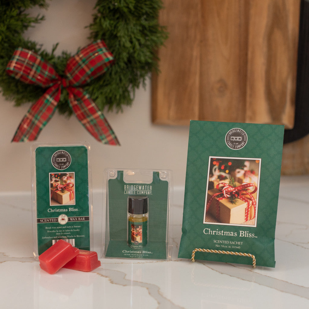 Three Christmas Bliss Flameless Bundle products by Bridgewater Candle Company, in green packaging, are on a counter with red wax melts, a wreath with plaid bow, and a wooden board in the background.