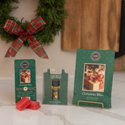 Three Christmas Bliss Flameless Bundle products by Bridgewater Candle Company, in green packaging, are on a counter with red wax melts, a wreath with plaid bow, and a wooden board in the background.