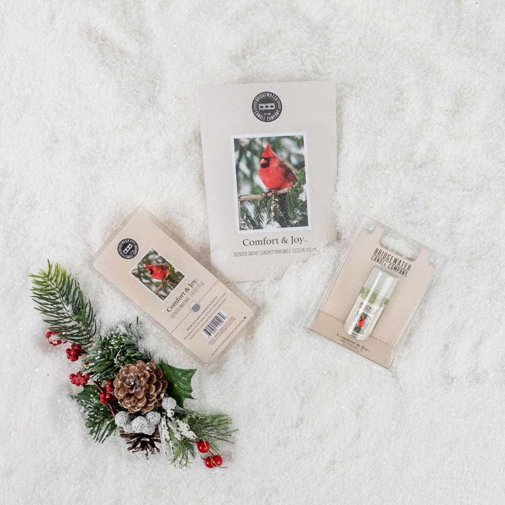 A flat lay of Bridgewater Candle Companys Comfort & Joy Flameless Bundle on white snow, featuring a candle, lip balm, cardinal photo card, pinecone, berries, evergreen sprig decor, and a home fragrance oil for festive cheer.