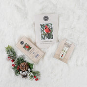A flat lay of Bridgewater Candle Companys Comfort & Joy Flameless Bundle on white snow, featuring a candle, lip balm, cardinal photo card, pinecone, berries, evergreen sprig decor, and a home fragrance oil for festive cheer.