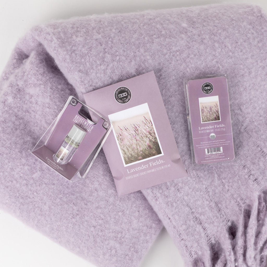 The Lavender Fields Flameless Bundle by Bridgewater Candle Company features a soft, light purple blanket with three matching lavender-packaged items—a vial, packet, and box—ideal for creating a calming home fragrance.