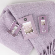 The Lavender Fields Flameless Bundle by Bridgewater Candle Company features a soft, light purple blanket with three matching lavender-packaged items—a vial, packet, and box—ideal for creating a calming home fragrance.