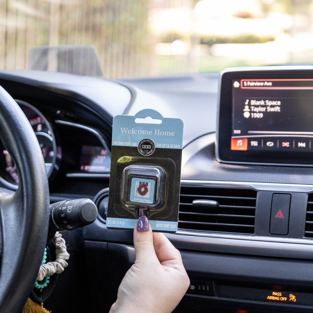 A person holds a Bridgewater Candle Company Welcome Home Auto Vent Clip in a car, with “Blank Space” by Taylor Swift on the dashboard screen and the steering wheel and instrument panel in view—enjoy fresh fragrance as you drive.