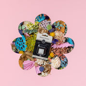 The Bridgewater Candle Company Sweet Grace Auto Vent Clip, in sleek black packaging, sits on a bed of colorful dried flowers against a pink background—ideal for fans of auto vent fragrances.