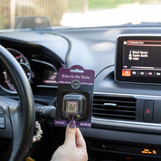 A person holds a Bridgewater Candle Company Kiss in the Rain Auto Vent Clip inside a car as sunlight streams through the windshield and Blank Space by Taylor Swift plays on the dashboard.