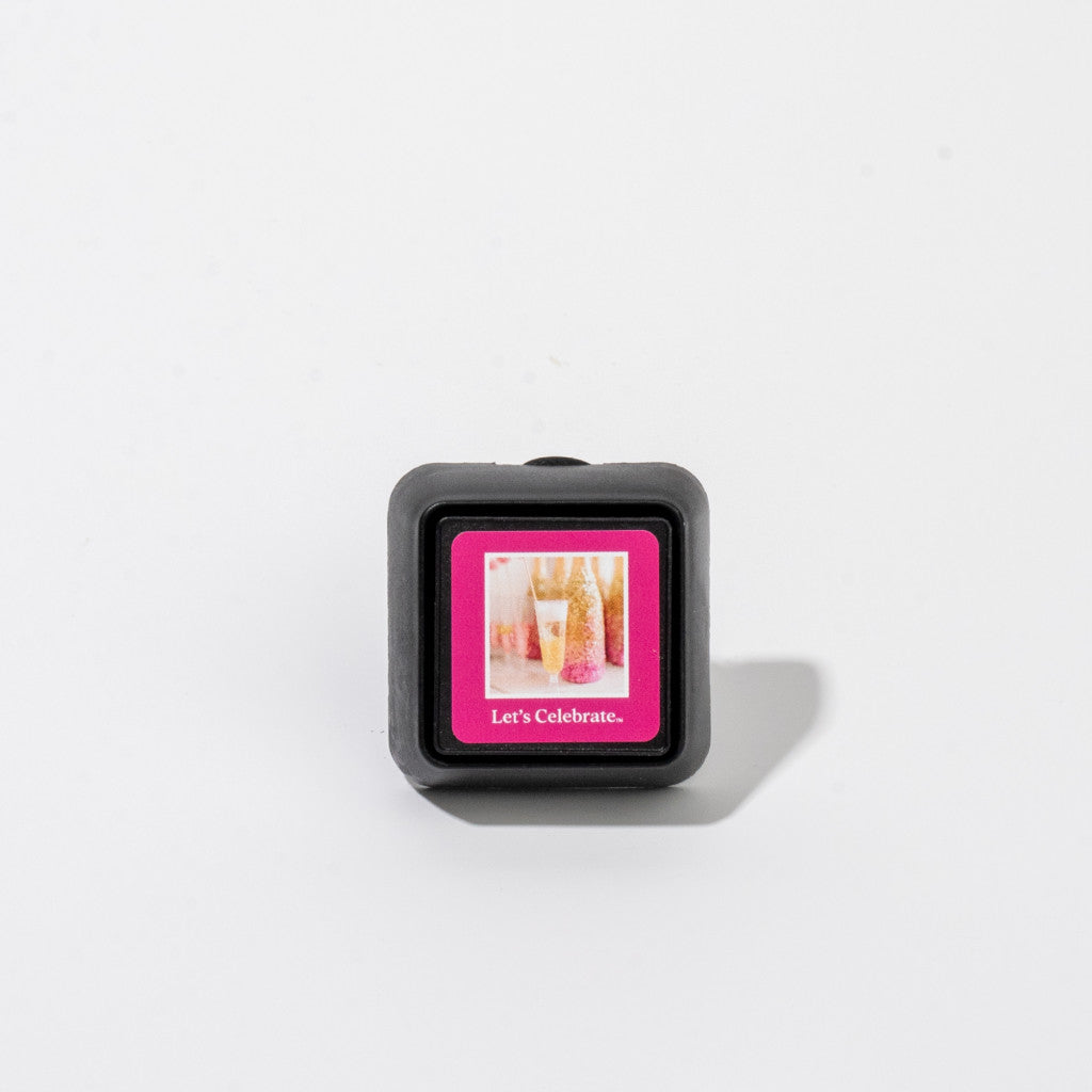 The Let’s Celebrate Auto Vent Clip by Bridgewater Candle Company is a small, square black vent clip with a pink label showing two champagne glasses and the text “Lets Celebrate.”—ideal for adding a festive touch to your car.