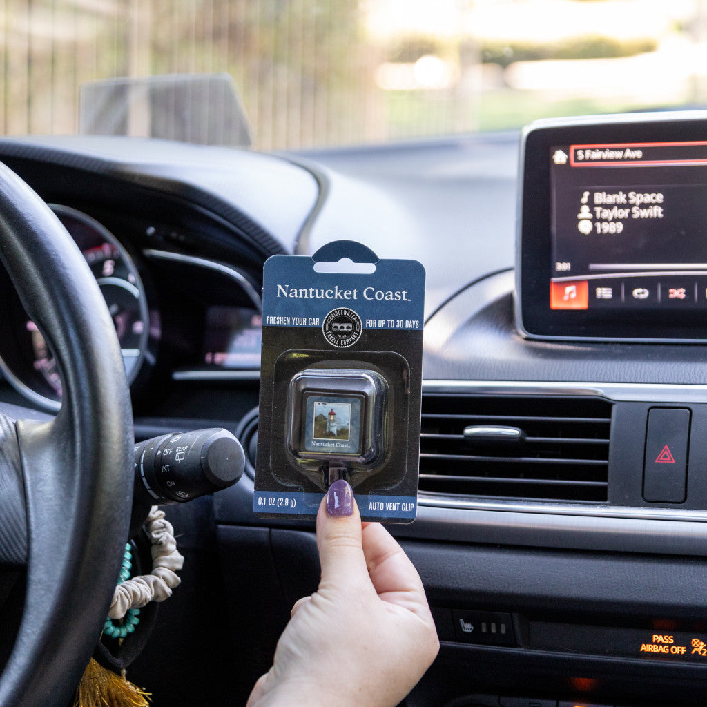 A hand holds a Bridgewater Candle Company Nantucket Coast Auto Vent Clip inside a car, with the dashboard, steering wheel, and a music display featuring Blank Space by Taylor Swift visible in the background.