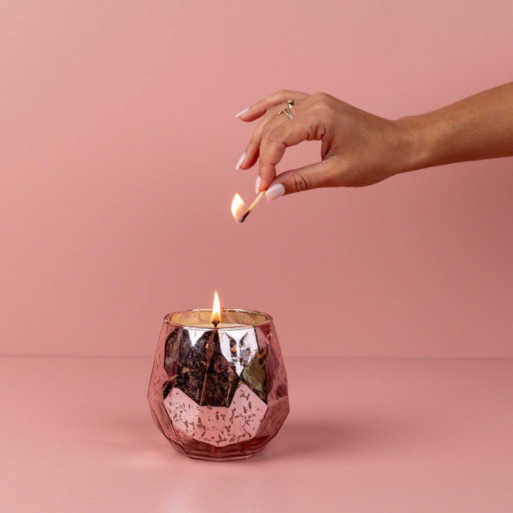 A hand with manicured nails lights the Bridgewater Candle Company Sweet Grace Collection Candle #011, showcased in a faceted metallic pink holder against a matching background, highlighting its best-selling fragrance and premium soy-blend wax.