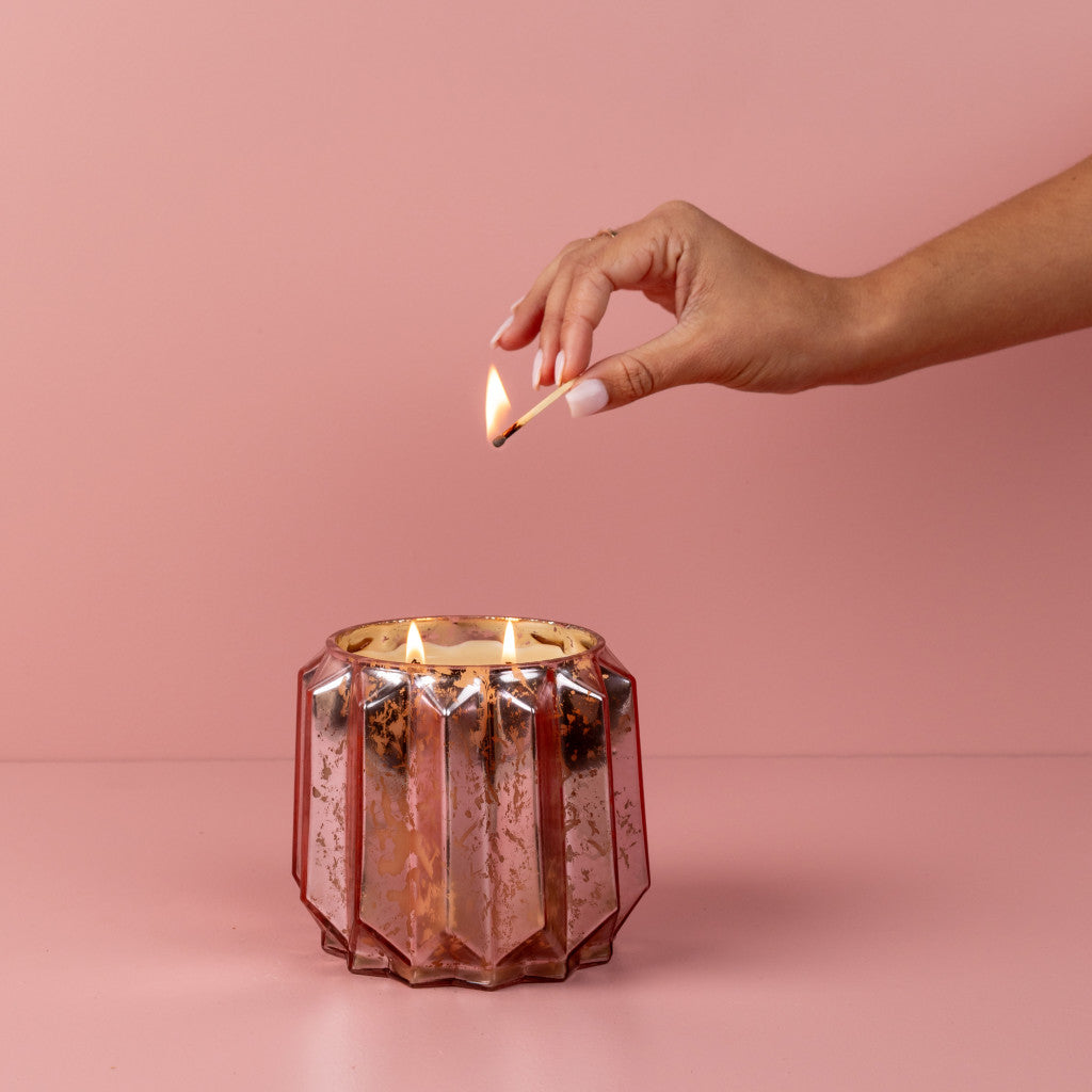 A hand with manicured nails lights a match to ignite the Bridgewater Candle Company Sweet Grace Collection Candle #021, its pink metallic finish glowing with three flames against a pink background.