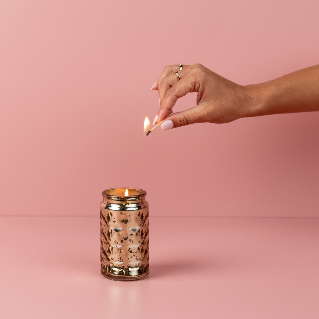 A manicured hand with a ring lights the Bridgewater Candle Company Sweet Grace Collection Candle #022, displayed in a gold-patterned glass jar with premium fragrance and soy-blend wax against a solid pink background.
