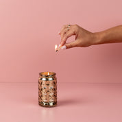 A manicured hand with a ring lights the Bridgewater Candle Company Sweet Grace Collection Candle #022, displayed in a gold-patterned glass jar with premium fragrance and soy-blend wax against a solid pink background.