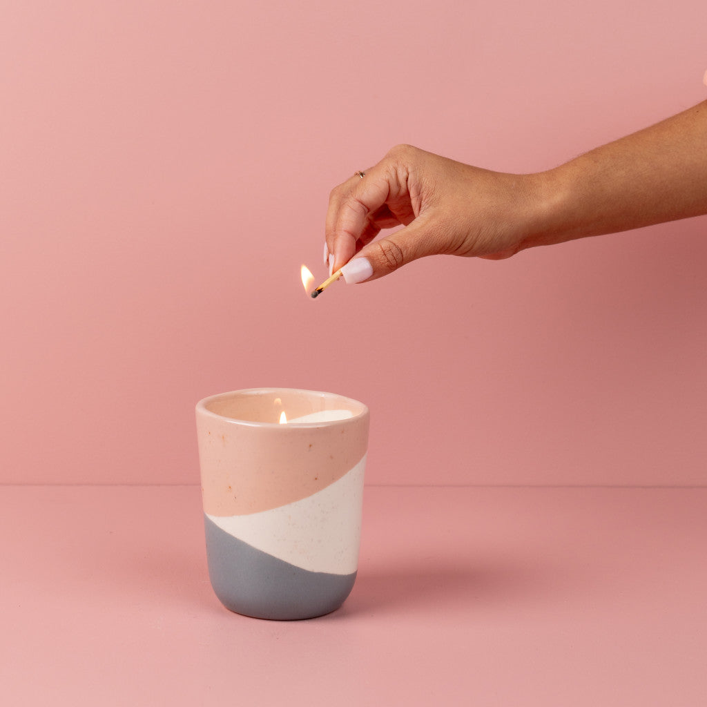 A hand with painted nails lights the Bridgewater Candle Company Sweet Grace Collection Candle #041, made with soy-blend wax, in a pink and gray ceramic holder against a pink background.