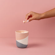 A hand with painted nails lights the Bridgewater Candle Company Sweet Grace Collection Candle #041, made with soy-blend wax, in a pink and gray ceramic holder against a pink background.