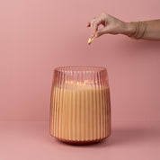A hand with bracelets lights the Sweet Grace Collection Candle #043 by Bridgewater Candle Company, featuring five cotton wicks and soy-blend wax in a ribbed rose glass holder, set against a pink background.