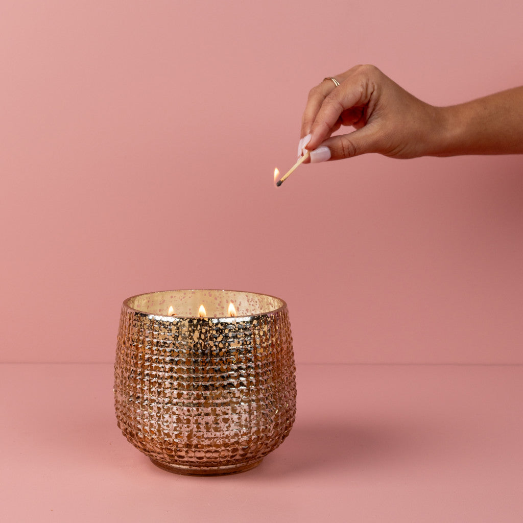 A hand with painted nails lights Bridgewater Candle Companys Sweet Grace Collection Candle #051, a large, textured metallic soy-blend candle, with a match against a pink background.